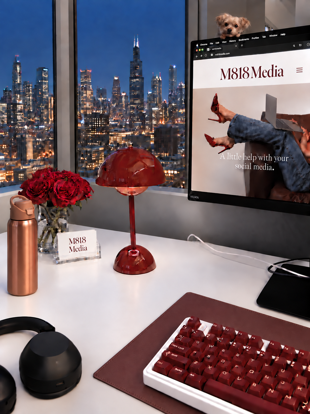 M818 Media desk with red roses, a copper water bottle, M818 Media business cards, a small Yorkie perched on the monitor, and the Chicago skyline glowing at night